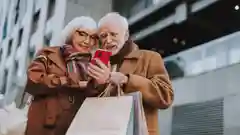 An elderly couple wearing coats standing outdoors with shopping bags, smiling together while looking at a red smartphone.