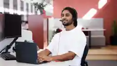 A young man sits at an office desk, smiling while working on a laptop, in a white t-shirt and a black over-ear headset with a microphone.