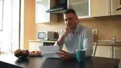 A man sitting at a kitchen table looking concerned while holding his electricity bill.
