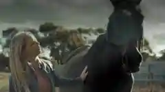 A woman in a denim jacket standing outdoors and petting a large black horse on a farm.