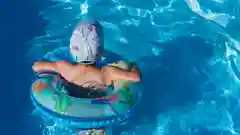 A child wearing a sun hat floating in a bright blue swimming pool in an inflatable ring.