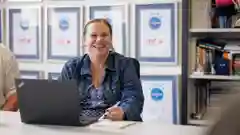 A woman sits at a white table, smiling warmly at the camera. An open laptop sits on the table in front of her.