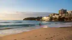 A person carrying a surfboard walks along a sandy beach towards the ocean, with coastal buildings in the background.