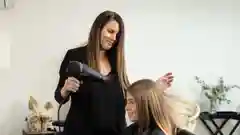 A hairstylist in a black top blow-dries and styles a seated client's long blonde hair in a modern, bright salon with minimalist white decor.