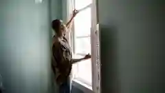 A man stands inside a home, opening or inspecting a double-glazed window next to white wooden shutters