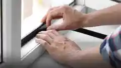Hands applying adhesive weather stripping to a window frame for draught proofing.