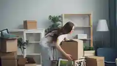 A woman lifting a cardboard box in a living room filled with packed belongings and indoor plants.