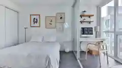 A bright, modern apartment bedroom with a white bed, desk, and large windows.