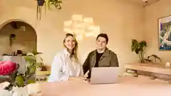 A smiling couple sits at a pink table with a laptop in a warm ceramics studio, a neon sign is glowing behind them.