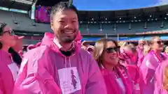 A smiling man in a pink rain poncho stands among a crowd of people also wearing pink ponchos inside a large open-air stadium on a sunny day.