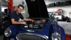 A smiling mechanic in a navy polo leans over the open hood of a classic blue sports car in a well-equipped automotive workshop.