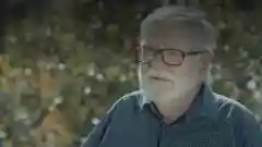 An older man with a grey beard and glasses looking off-camera outdoors.