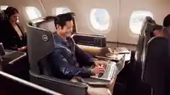 A man in a blue blazer works on a laptop in a business class airline seat with a drink and snack on the tray, cabin windows in the background."