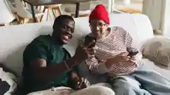 Two friends sitting on a couch, smiling as one shows something on his smartphone screen to the other.
