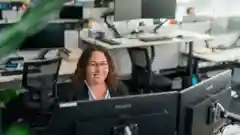 A smiling woman with curly hair and glasses sits at a desk with dual monitors in a busy open-plan office with workstations behind her.