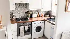 A white front-loading washing machine installed under a kitchen bench next to a stainless steel oven