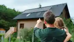 A father holding his young daughter points toward their wooden house with rooftop solar panels, seen from behind against a green hillside."