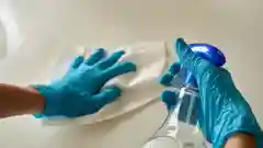 A person wearing blue rubber gloves wiping down a surface with a paper towel and a spray bottle.