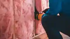 A person installing pink fiberglass insulation batts into a wooden wall frame.