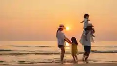 At sunset, a family of four strolls through shallow ocean water, with young children perched on their parents’ shoulders beneath a glowing orange sky.