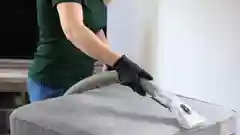 A person wearing a dark green shirt and black gloves vacuums a grey upholstered ottoman.