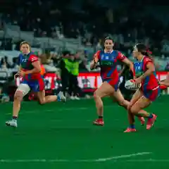 Three female rugby league players in red and blue jerseys running on a lit stadium field at night, one carrying the ball.