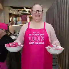  A smiling woman wearing glasses and a bright pink BCNA apron that reads "Buy a fun bun, 100% goes to BCNA" holds two bowls of pink buns in a kitchen.