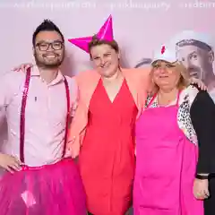  Three people pose together at a pink-themed BCNA  event, all dressed in pink outfits including a tutu, red dress, and pink apron with hat.