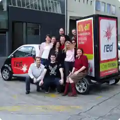 A group of about ten people posing together in a parking area beside a small branded red car and a promotional trailer.