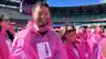 A smiling man in a pink rain poncho stands among a crowd of people also wearing pink ponchos inside a large open-air stadium on a sunny day.