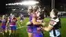 Female rugby players in blue and red jerseys celebrate together on the field at night.