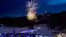 A large fireworks display lights up the night sky above a packed snow resort, with colourful lights illuminating the crowd below.