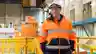 Smiling worker in orange hi-vis jacket and hard hat standing confidently inside a large hydroelectric power station