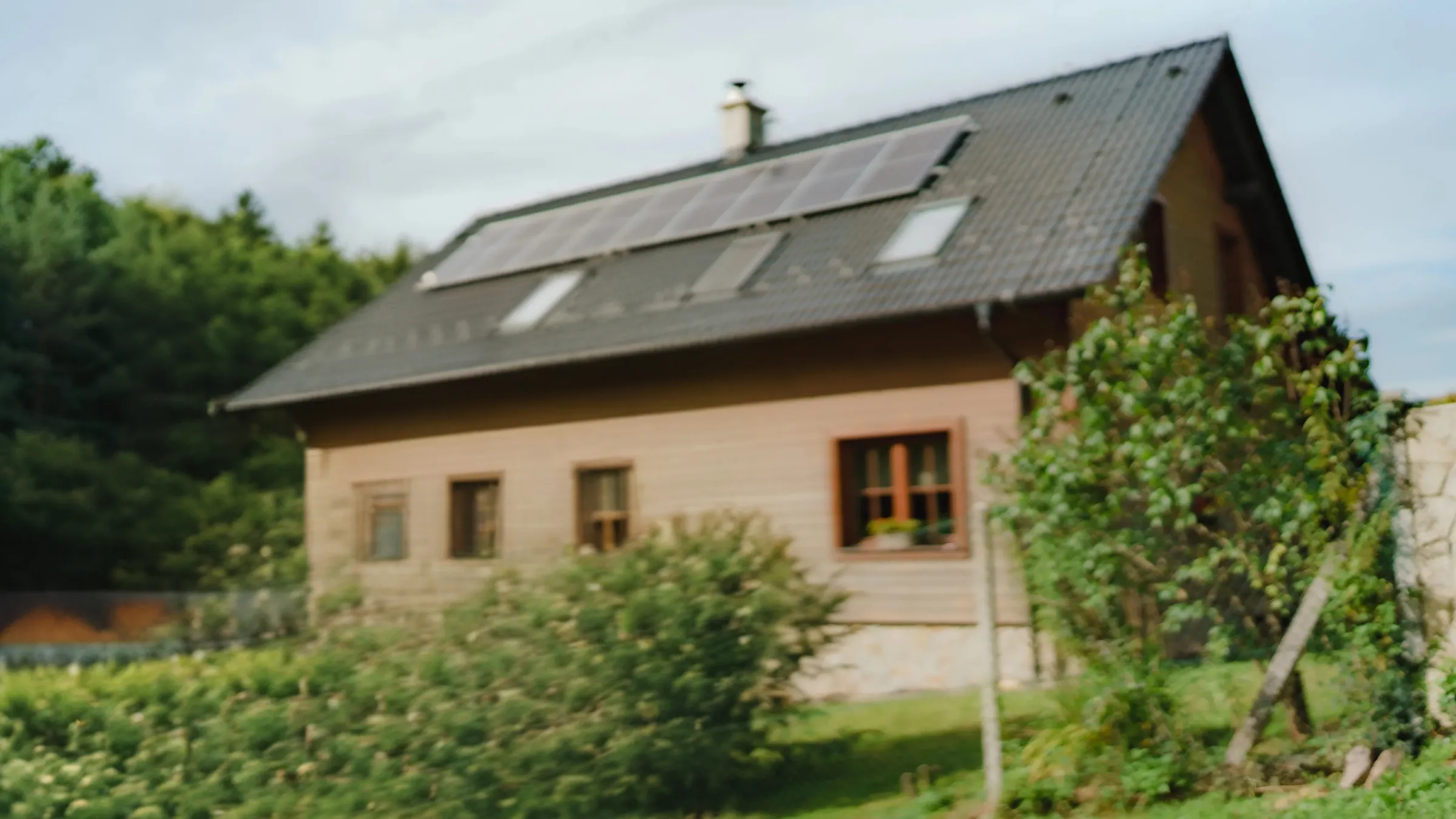 A smiling couple embraces in their garden in front of a wooden house with solar panels on the roof, surrounded by lush green landscaping.