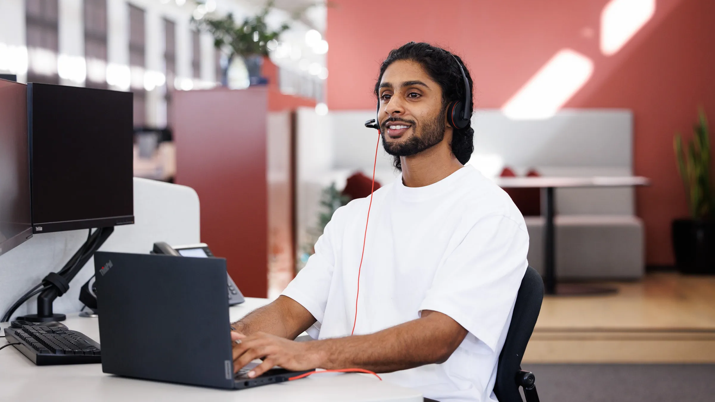 A young man sits at an office desk, smiling while working on a laptop, in a white t-shirt and a black over-ear headset with a microphone.