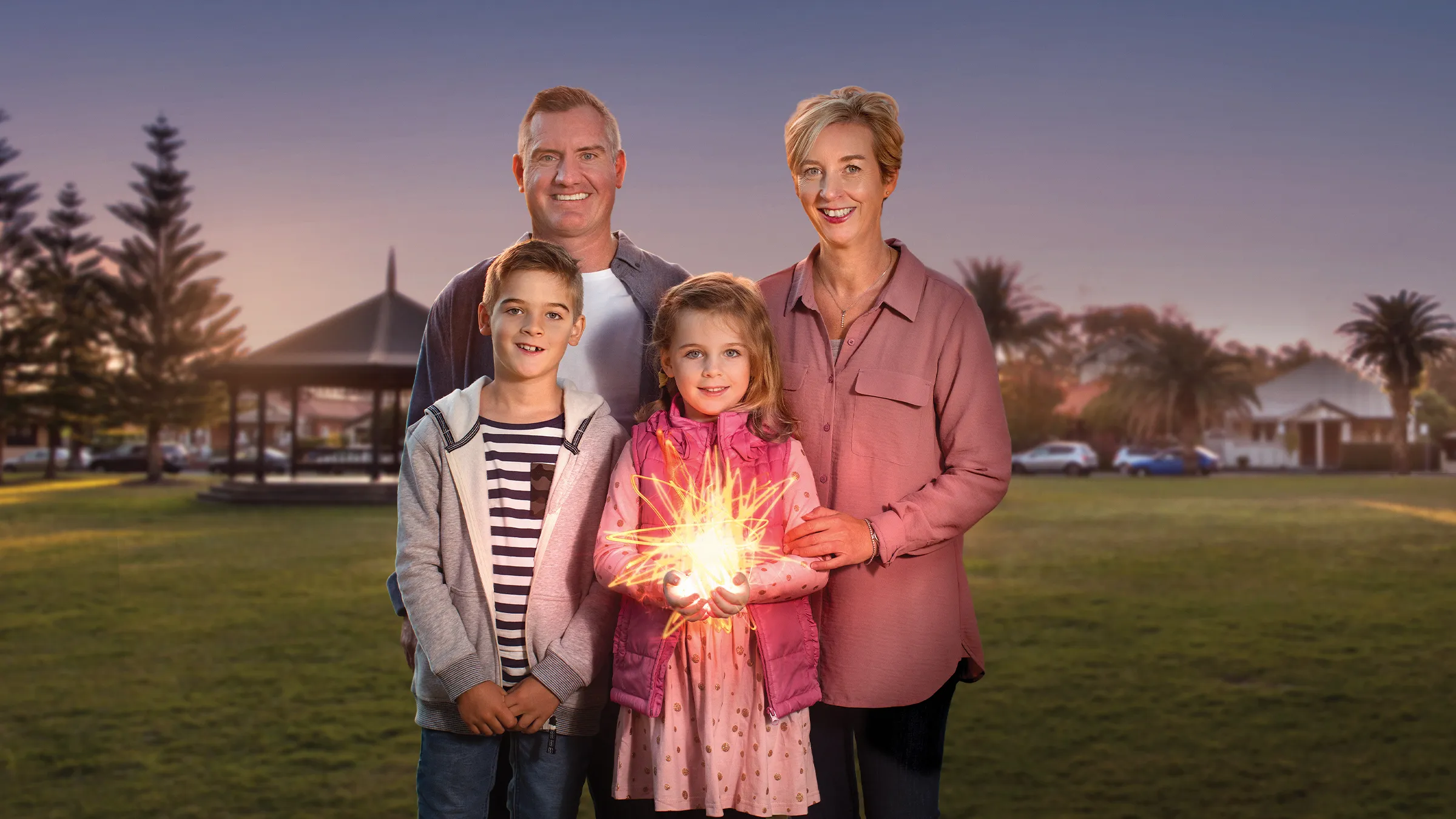 A smiling family of four stands outdoors in a park at dusk, with the young daughter holding a glowing, starburst-like light in her hands.