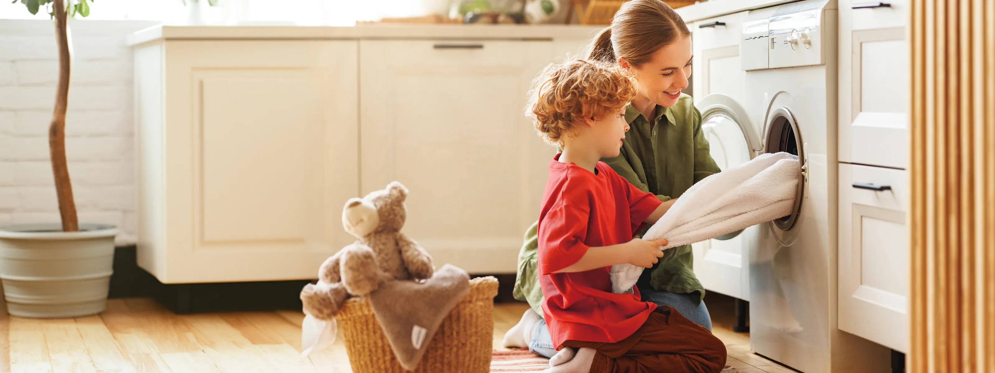 A mother and young child kneel beside a washing machine, loading white laundry together in a bright, cozy utility room with a woven teddy bear nearby.
