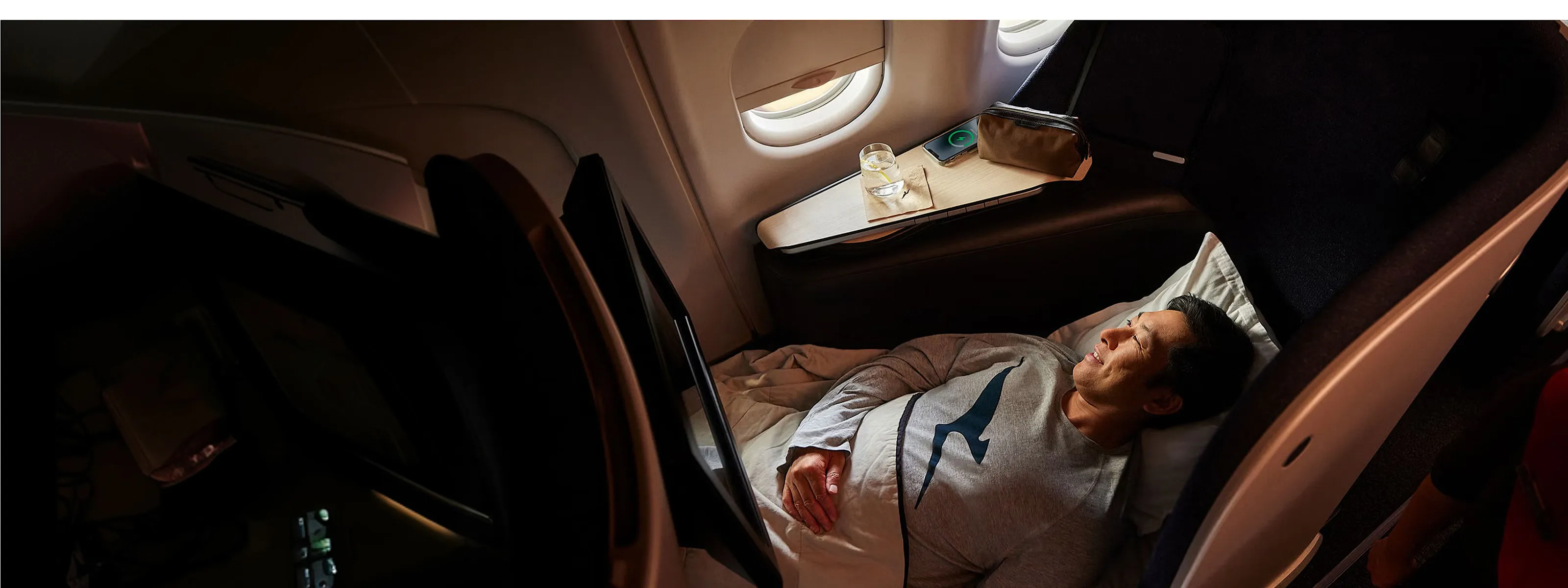 A man sleeps peacefully in Qantas business class. He wears Qantas branded pyjamas as he relaxes with the light from an open window on his face.
