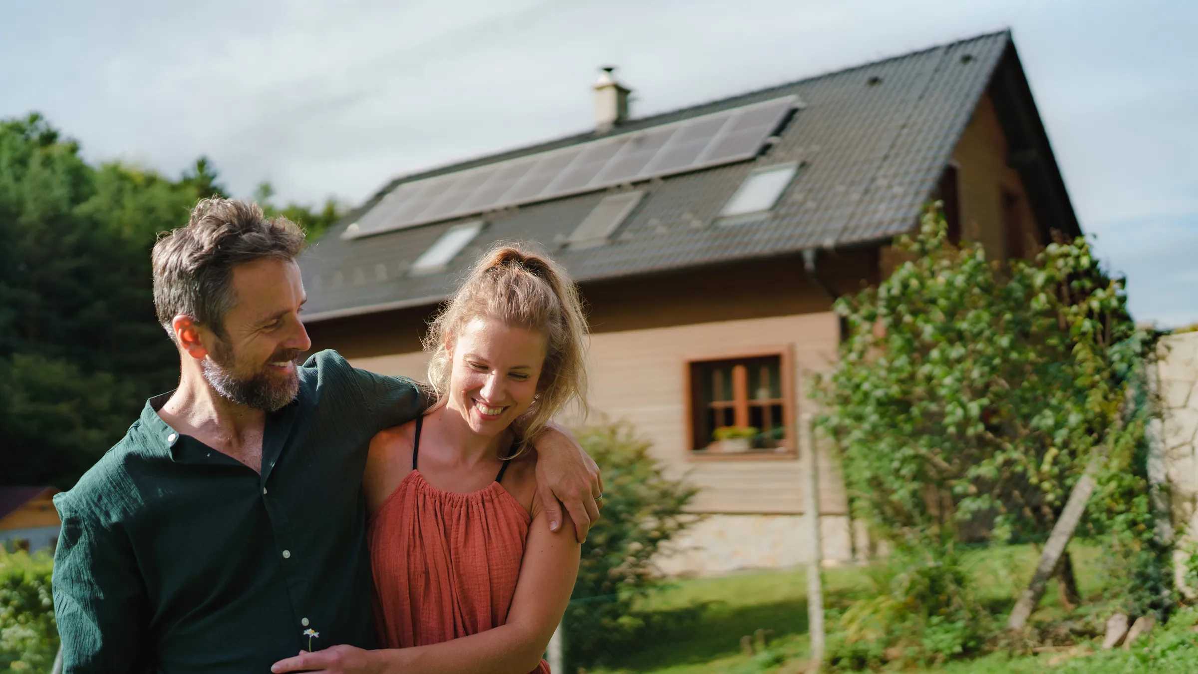 A smiling couple embraces in their garden in front of a wooden house with solar panels on the roof, surrounded by lush green landscaping.