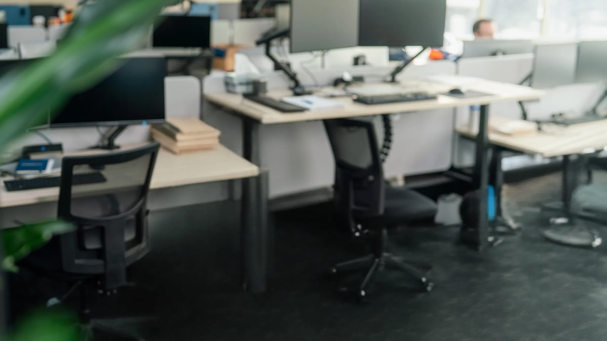 A smiling woman with curly hair and glasses sits at a desk with dual monitors in a busy open-plan office with workstations behind her.