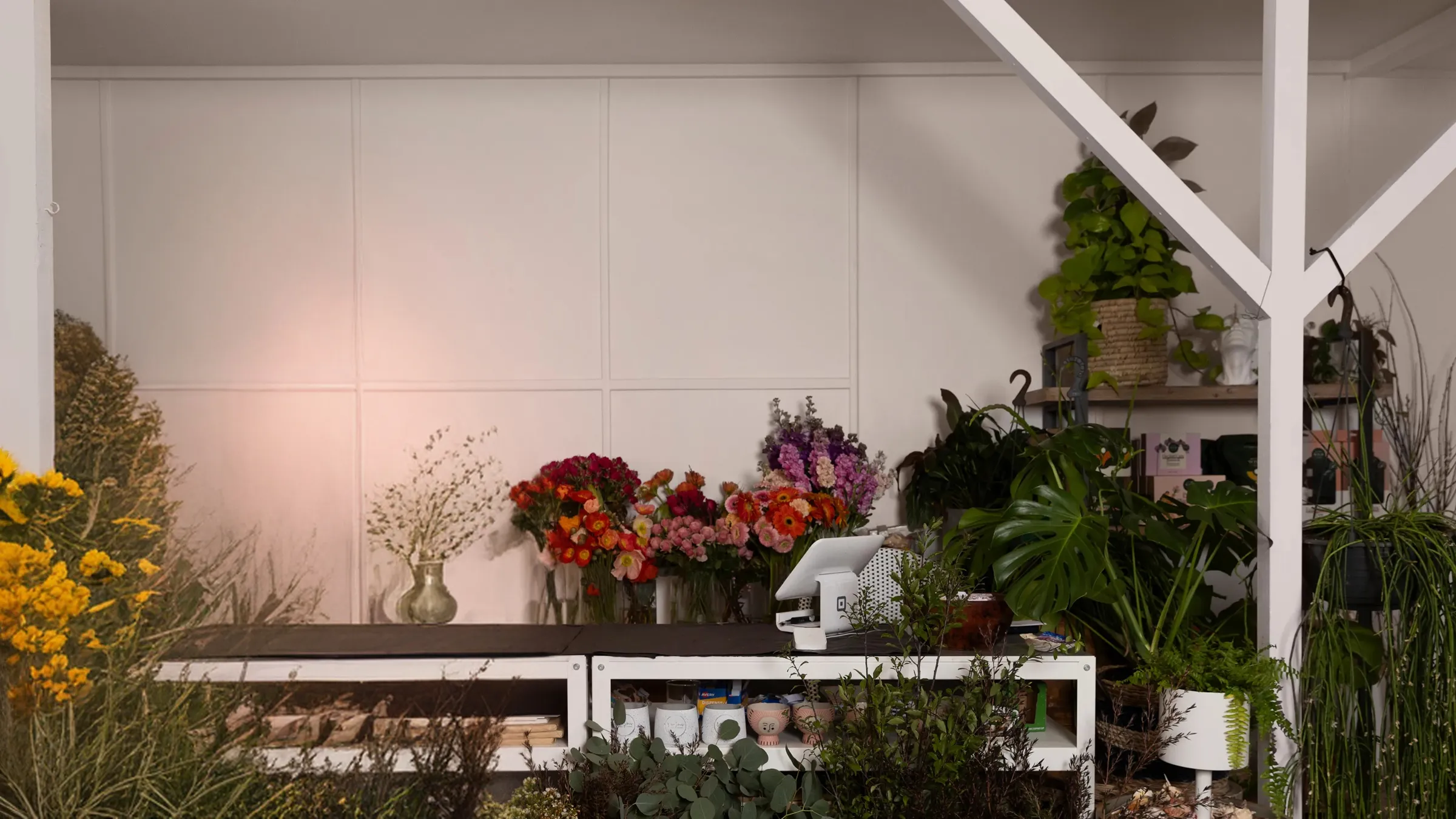 A florist in a black apron standing behind a counter in her shop, surrounded by green plants, flowers, and decorative wall ornaments.