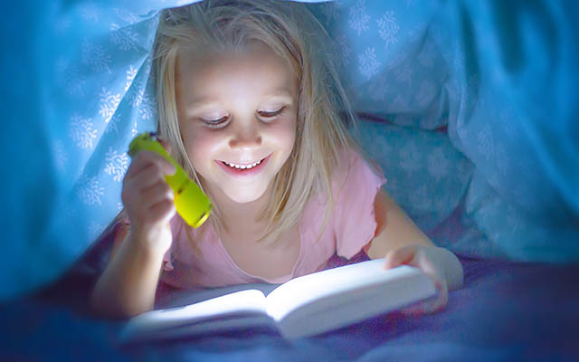 Little girl lying under bed covers reading book in the dark with torch